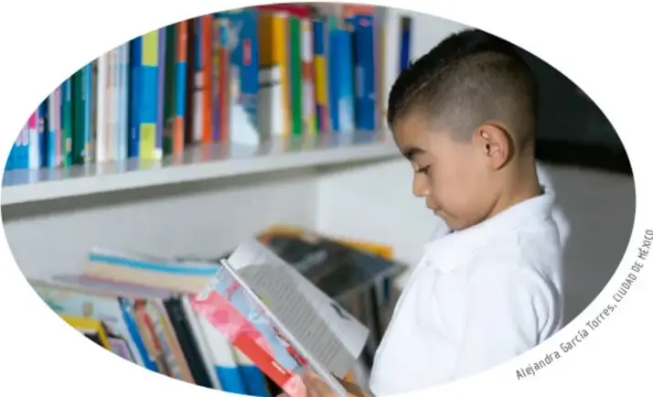 Fotografía ovalada de un niño de camisa blanca leyendo de pie frente a una estantería con libros de colores; crédito de la Alcaldía García Torres, Ciudad de México.