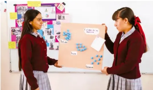 Fotografía de dos estudiantes de telesecundaria con uniforme (chamarra roja) presentando un cartel de cartón con etiquetas (Señal, Luz, Sonido, Imágenes) y fichas adhesivas frente a una pared con trabajos escolares.