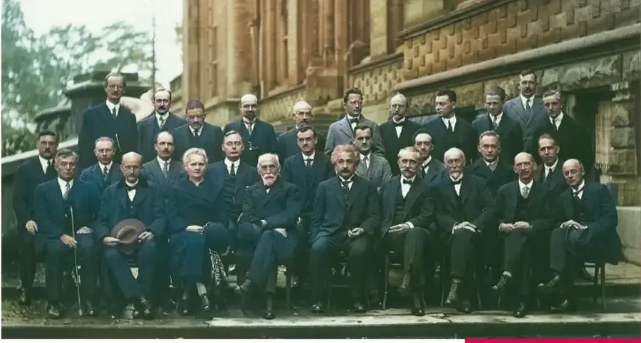 Fotografía antigua en color de una reunión internacional de científicos: un numeroso grupo de hombres vestidos de traje posa frente a un edificio de piedra, con Marie Curie entre ellos.