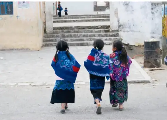 Tres niñas de espaldas caminando por un patio escolar vistiendo blusas y faldas bordadas de colores (azul, morado, verde).