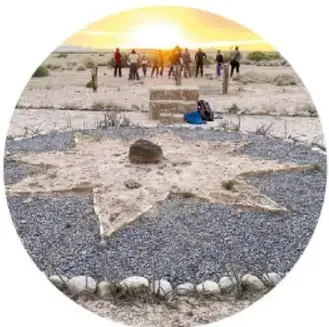 Fotografía circular del atardecer en la zona del silencio: personas junto a una estructura de piedras en el desierto, con un marcador en el suelo formando una estrella o rayo con piedras blancas y oscuras.