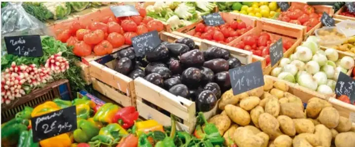 Puesto de mercado con verduras y frutas: jitomates, berenjenas, cebollas, papas, chiles y rábanos, con etiquetas de precio.