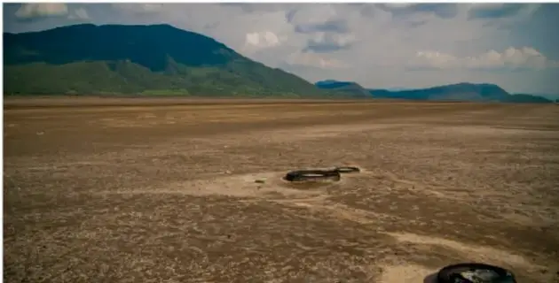 Fotografía de un paisaje árido con un espejo o cuerpo de agua seco en primer plano, suelo desnudo y montañas verdes al fondo bajo un cielo con nubes, representando la escasez de agua.