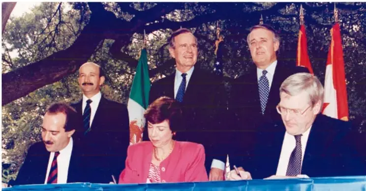 Fotografía de la firma del TLCAN con varios funcionarios, entre ellos Carlos Salinas de Gortari, y banderas de México, Estados Unidos y Canadá.