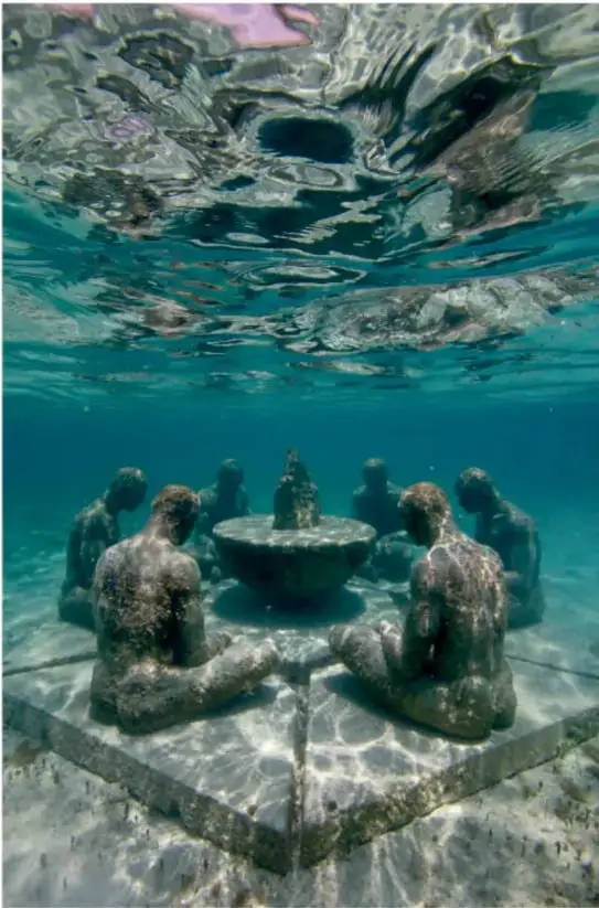 Fotografía subacuática de la escultura 'Entendimiento' de Elier Amado Gil. Muestra varias figuras humanas de piedra sentadas en círculo alrededor de una mesa circular, bajo el agua cristalina del mar de Quintana Roo. La luz solar se filtra desde la superficie.
