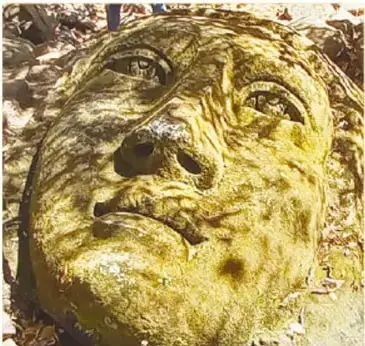 Fotografía cercana de una de las cabezas gigantes tallada en piedra cubierta de musgo, con rasgos de rostro indígena (ojos, nariz y boca labrados), entre rocas y árboles.