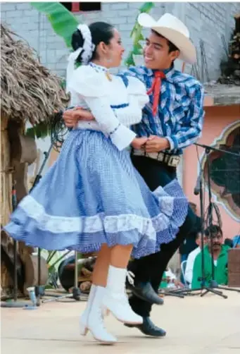 Fotografia de una pareja de bailarines de danza folclorica mexicana; la mujer viste blusa blanca, falda azul con olan y botas blancas, y el hombre camisa a cuadros, pantalon oscuro, sombrero y paliacate rojo. Fondo con techo de palma y publico.