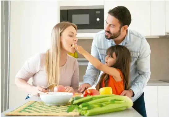 Fotografía de una familia (madre, padre e hija) preparando alimentos frescos como apio y tomates en la cocina.