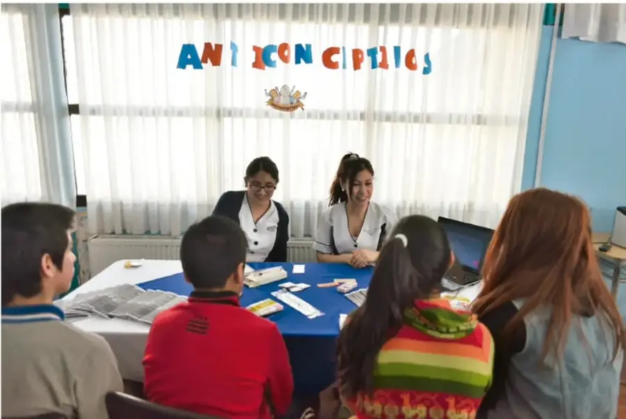Fotografía de una sesión informativa: dos profesionales atienden a un grupo de jóvenes alrededor de una mesa; en la cortina se leen letras de colores que forman la palabra 'ANTICONCEPTIVOS'.