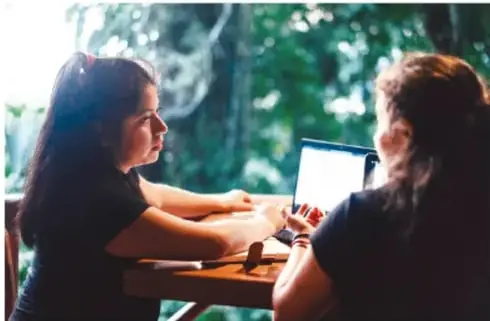 Dos adolescentes frente a una computadora portátil, una de ellas parece estar siendo aconsejada por la otra.