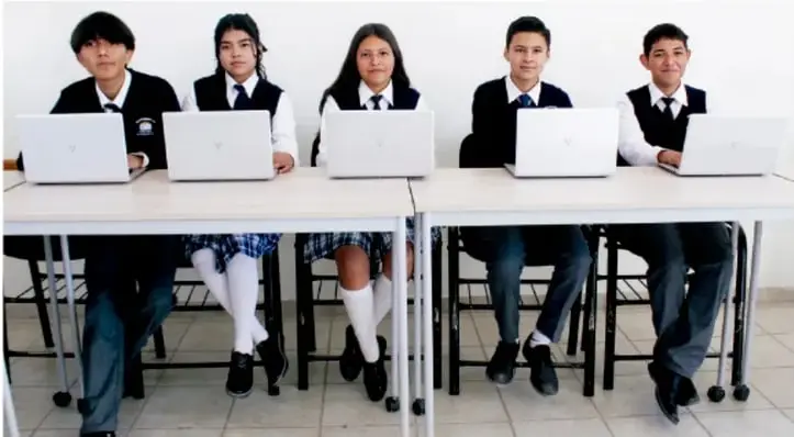 Fotografía de cinco estudiantes de telesecundaria con uniforme, sentados en dos mesas usando laptops (computadoras portátiles) blancas.