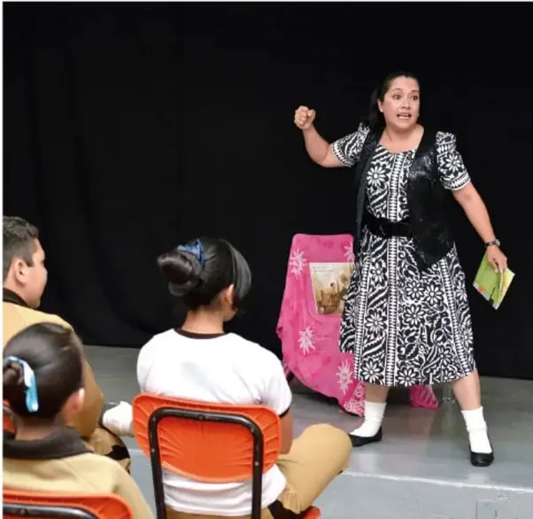 Fotografía de una actriz en escena interpretando ante un público infantil y juvenil sentado, aparentemente en una función teatral o de lectura dramatizada.