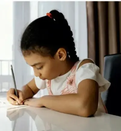 Fotografía de una niña escribiendo en su cuaderno, sentada en un escritorio junto a una ventana.