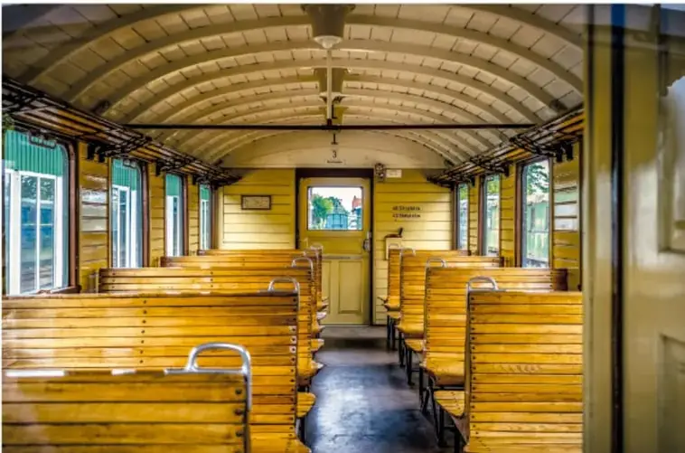 Interior de un vagón de pasajeros antiguo, con asientos de madera de color amarillo, techos curvos y lámparas colgantes.