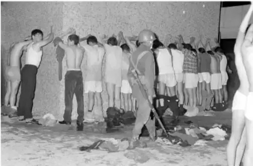 Fotografía en blanco y negro de personas detenidas y formadas contra una pared, vigiladas por un soldado armado.