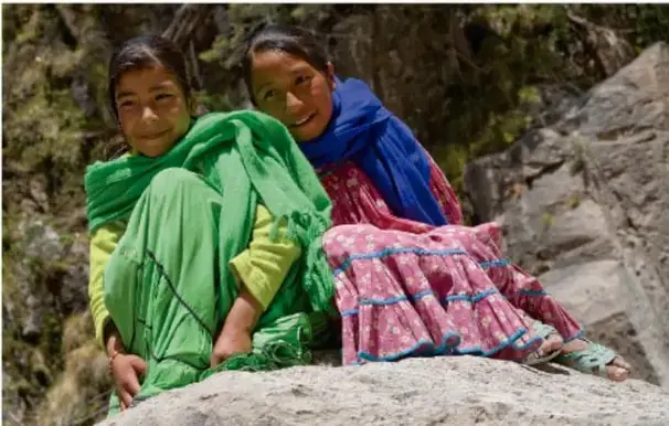 Fotografía de dos niñas indígenas vestidas con ropa tradicional de colores verde y rojo, sentadas sobre rocas en un entorno natural.