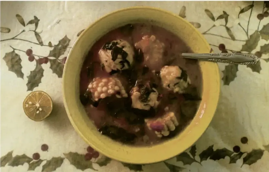 Fotografía cenital de un cuenco amarillo con una preparación tradicional en caldo rojizo que contiene granos, hierbas y piezas de carne o maíz; al lado hay medio limón, todo sobre un mantel con motivo de hojas de acebo y bayas rojas. Ilustra la idea de revivir el territorio "en sus comidas".