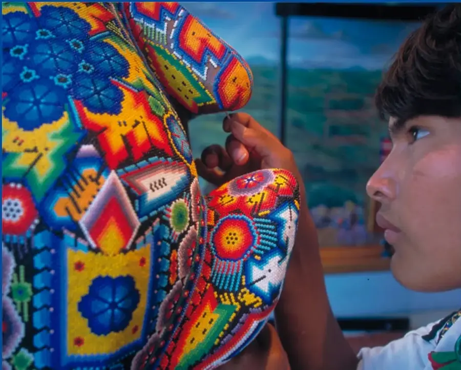 Fotografía de un estudiante de perfil observando y tocando una máscara tradicional mexicana altamente decorada con bordados y cuentas de colores.