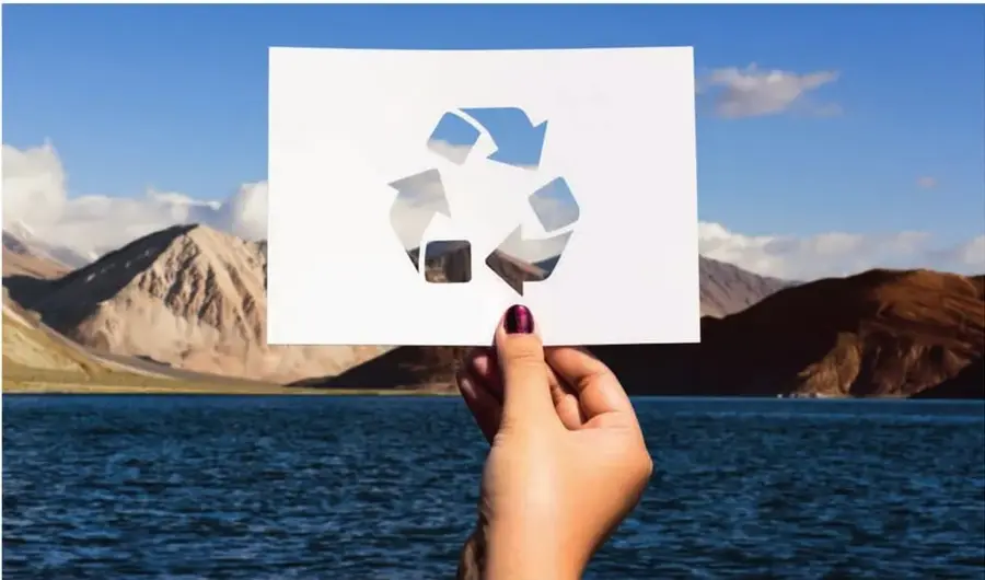 Fotografía de una mano sosteniendo papel recortado con el símbolo de reciclaje frente a un paisaje de montañas y lago.