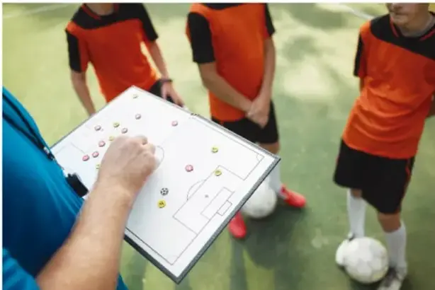 Un entrenador sostiene un pizarrón con la estrategia de un campo de fútbol y explica a tres jóvenes futbolistas con camisetas naranjas y pantalones negros durante una sesión de entrenamiento.