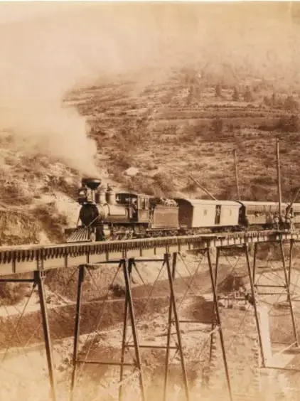 Fotografía antigua en blanco y negro de un tren de vapor cruzando un puente de madera, con montañas al fondo. Representa el ferrocarril como símbolo del progreso porfirista.