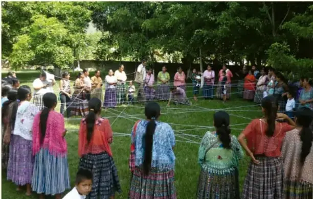 Fotografía de un grupo de personas, en su mayoría mujeres con ropa tradicional, formando un círculo en un parque mientras sostienen cuerdas o listones.