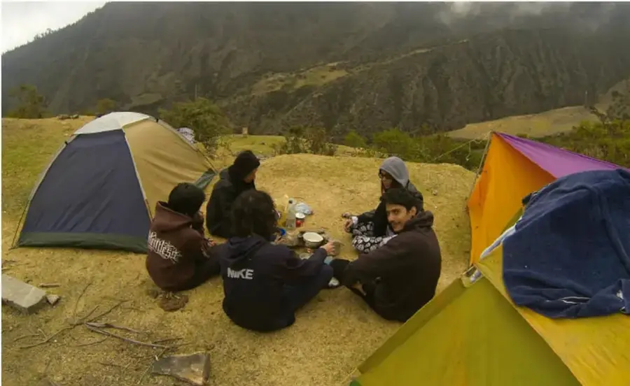 Fotografía de un grupo de jóvenes acampando en una zona boscosa de montaña: hay dos tiendas de campaña (una azul-verde y otra naranja-amarilla) y cinco personas sentadas en círculo alrededor de un fogón mientras conversan y preparan alimentos; el paisaje de fondo muestra montañas cubiertas de vegetación con neblina.