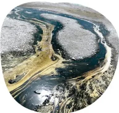 Derrame de petróleo formando charcos oscuros sobre el suelo junto a un río.