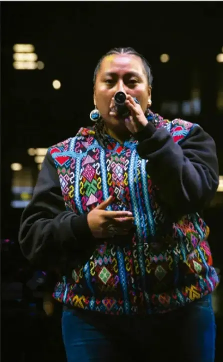 Fotografía de una rapera con chaleco bordado de colores, sosteniendo un micrófono durante una presentación en vivo.