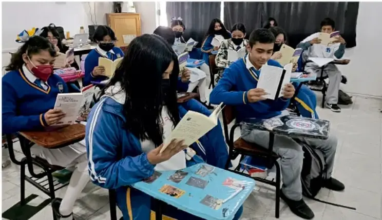 Fotografía de alumnos de Telesecundaria con uniforme azul y mascarillas, sentados en el aula leyendo libros y carpetas de trabajo. Crédito fotográfico: Luis Eduardo García Mérida, TIXLA.