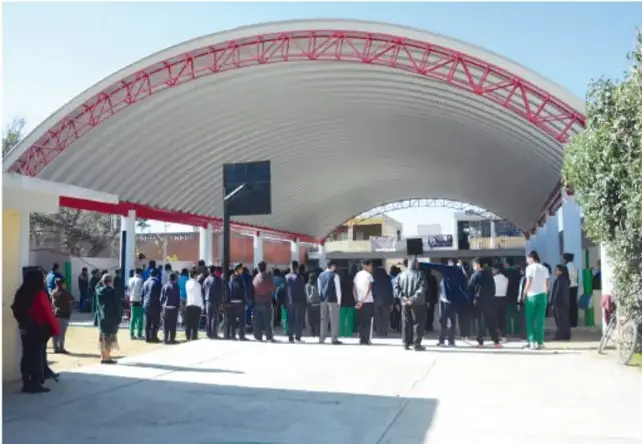 Patio escolar techado con estructura metálica curva y un grupo de estudiantes reunidos, posiblemente lugar de exhibición de la infografía.