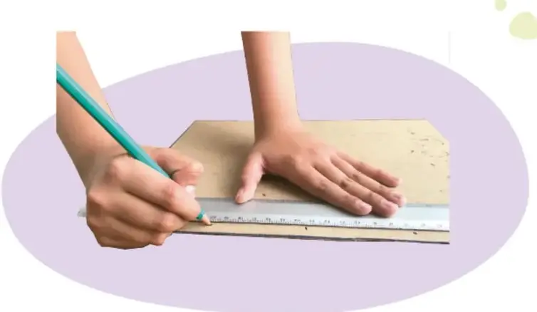 Manos de un estudiante sosteniendo una regla sobre un cartón y marcando con lápiz los puntos que se van a perforar.