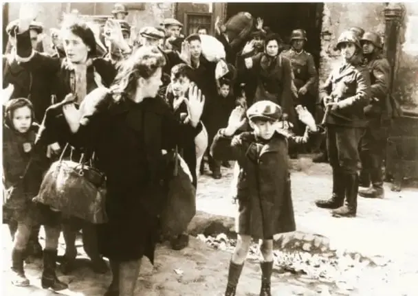 Fotografía histórica en blanco y negro de civiles (mujeres, niños y ancianos) con las manos en alto, custodiados por soldados con casco en una calle en ruinas; representa la población afectada por la ocupación durante la Segunda Guerra Mundial.