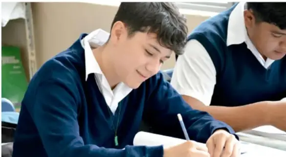 Fotografía de estudiantes con uniforme escolar (chamarra azul y camisa blanca) concentrados escribiendo en sus cuadernos durante una actividad de proyecto académico.
