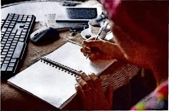 Persona escribiendo en un cuaderno junto a una computadora; representa el proceso de preparación para escribir.