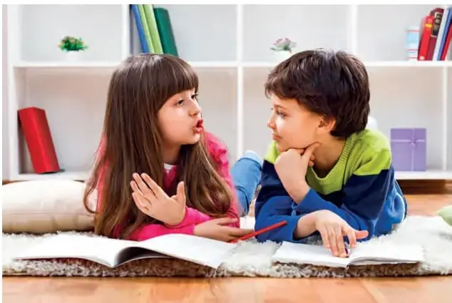 Fotografía de dos niños (una niña y un niño) recostados sobre una alfombra leyendo un libro abierto; al fondo, una estantería blanca con libros de colores.