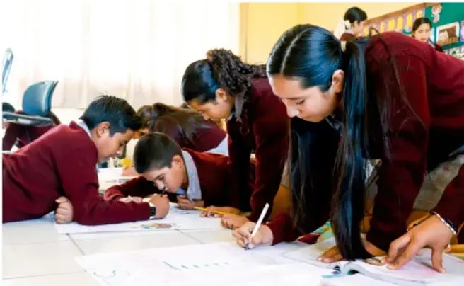 Fotografía de estudiantes en uniforme escolar trabajando en equipo, inclinados sobre una mesa escribiendo y dibujando en hojas de papel.