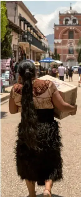 Fotografía de una mujer de espaldas con cabello largo trenzado, cargando una caja en una calle con comercios y un templo al fondo, ilustrando la vida comunitaria.