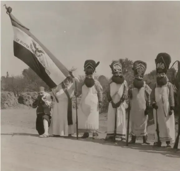 Fotografía en la que aparecen varios chinelos disfrazados con máscaras y vestimentas blancas, portando una bandera, en un camino al aire libre.