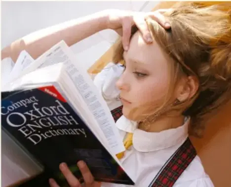 Fotografía de una estudiante rubia de uniforme escolar hojeando un diccionario Oxford English Dictionary; apoya la cabeza con una mano en actitud de estudio.