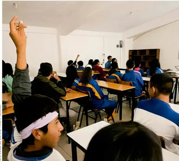 Fotografía de un aula de telesecundaria con estudiantes sentados en sus pupitres, algunos levantando la mano, participando en clase. Crédito: Luis Eduardo García Méndez, Tlaxiaca.