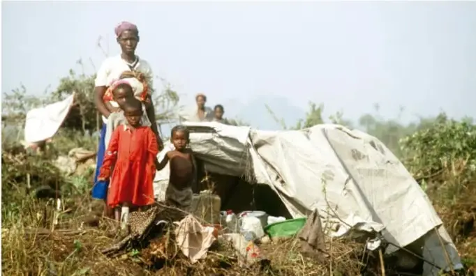 Fotografía a color de una familia desplazada junto a una refugio improvisado de lona plástica en campo abierto: una mujer con un niño cargado y dos niños de pie entre arbustos y utensilios.