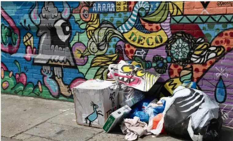 Mural de graffiti muy colorido con figuras, letras y colores llamativos, frente al cual hay bolsas de basura negras, cajas de cartón y residuos desechados en el piso, ilustrando el concepto de arte basura.