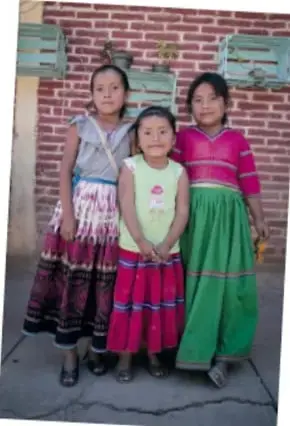 Tres niñas de pie frente a un muro de ladrillo, con ropa tradicional de colores (faldas verde y rosa).