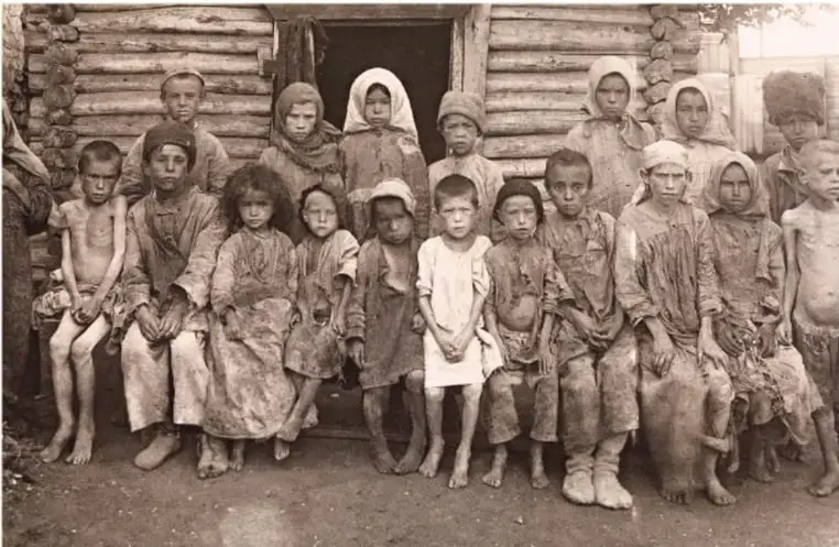 Fotografía en blanco y negro de un grupo numeroso de campesinos rusos, en su mayoría niños y mujeres, con ropas humildes, frente a una construcción de madera. Refleja la pobreza y las condiciones de vida del campesinado ruso a principios del siglo XX.
