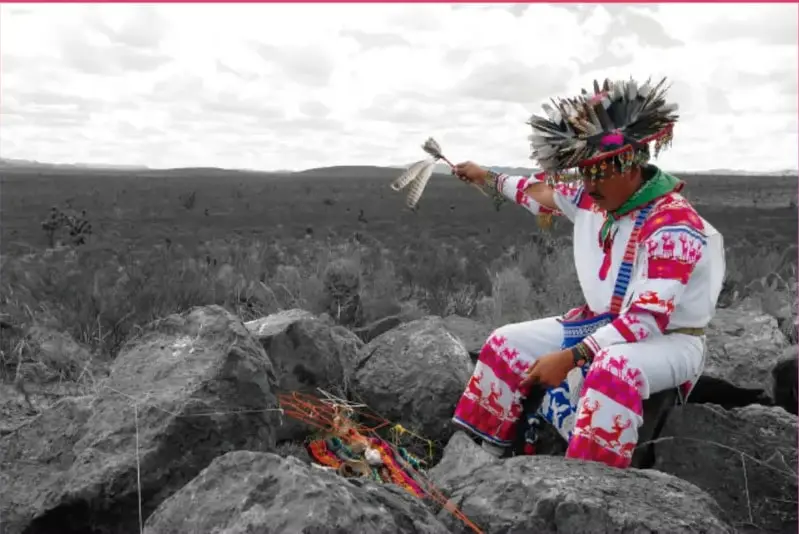 Fotografía en blanco y negro con detalles en color: persona con vestimenta tradicional emplumada, sentada entre rocas en un paisaje abierto, sosteniendo un instrumento ceremonial; crédito a Salatiel Barragán Santos, Veracruz.