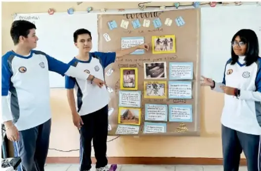 Fotografía de tres estudiantes de telesecundaria frente a un mural o cartelera de trabajo colaborativo del proyecto, señalando distintos elementos del panel.