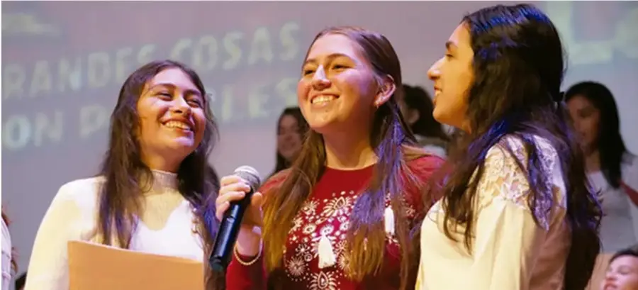 Fotografía de tres jóvenes sonrientes en un evento comunitario; una de ellas sostiene un micrófono. Detrás se lee el lema 'Grandes cosas son posibles'.