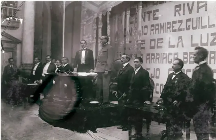 Fotografía en blanco y negro de un acto oficial con Venustiano Carranza y varios funcionarios; algunos de pie y otros sentados, con vestimenta formal de la época.