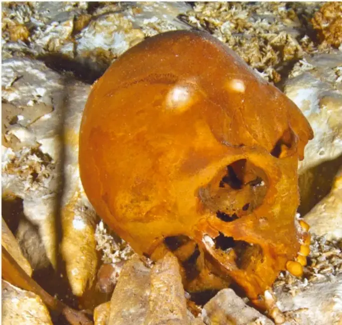 Fotografía subacuática del cráneo de Naia en el fondo de la caverna inundada, de color ámbar, entre rocas y sedimentos.
